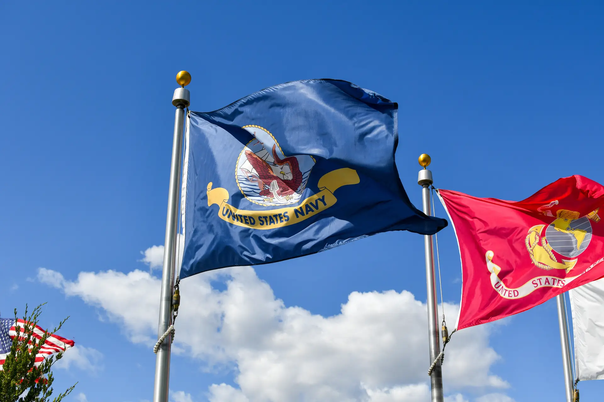 U.S. Navy flag and Marine Corps flag waving on flagpoles against a blue sky with white clouds.