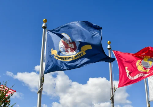 U.S. Navy flag and Marine Corps flag waving on flagpoles against a blue sky with white clouds.